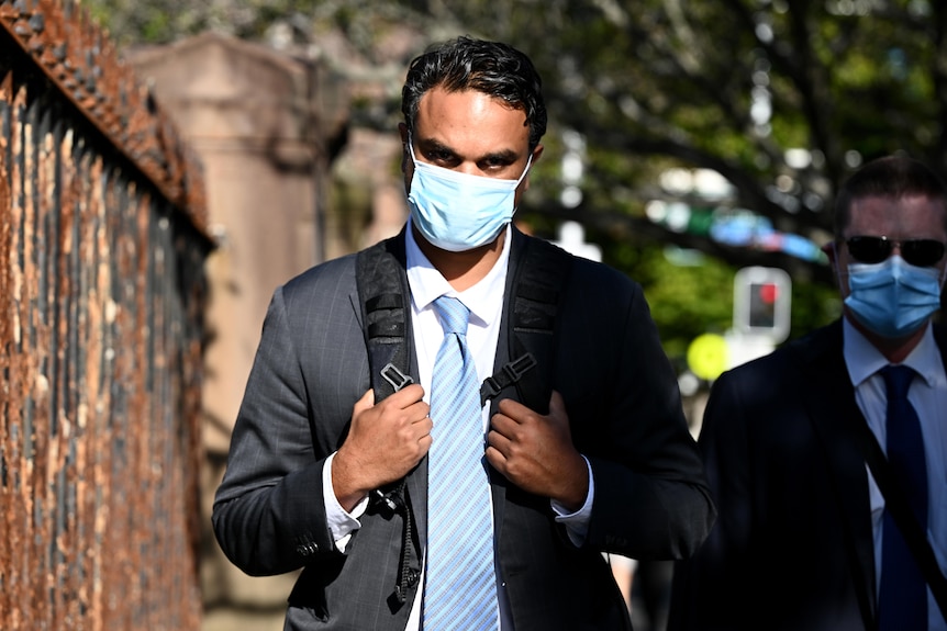 A male wearing a surgical mask walks past a metal fence holding the straps of his backpack