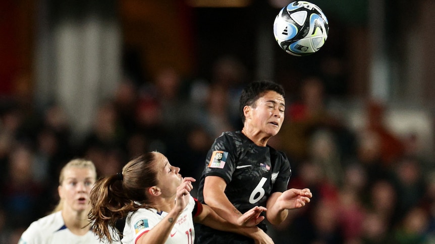 A New Zealand player in black head butts a white ball while fending off a Norwegian player dressed in white.