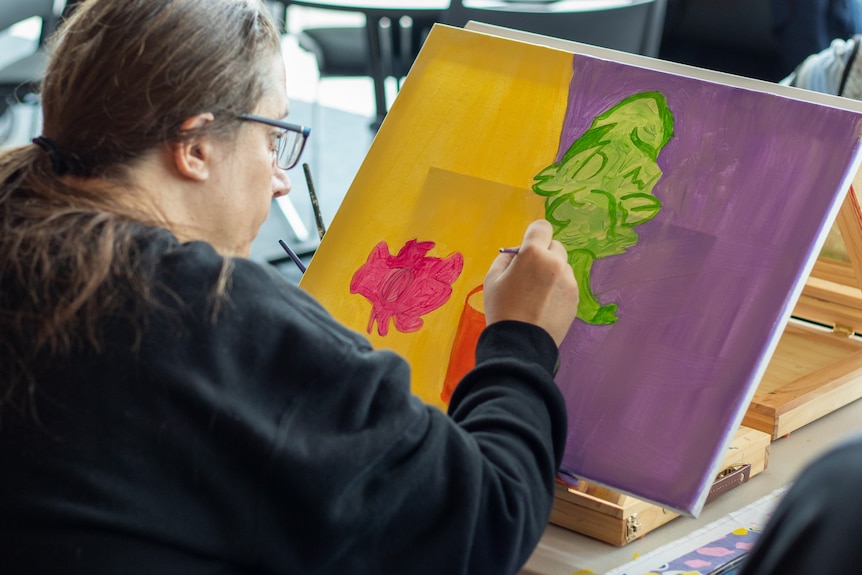 A woman is leaning over a purple and yellow canvas, she is painting a green plant in the centre
