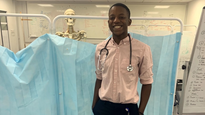 A dark-skinned young man standing in a medical teaching room with skeleton behind him. He is smiling and wearing a stethoscope.