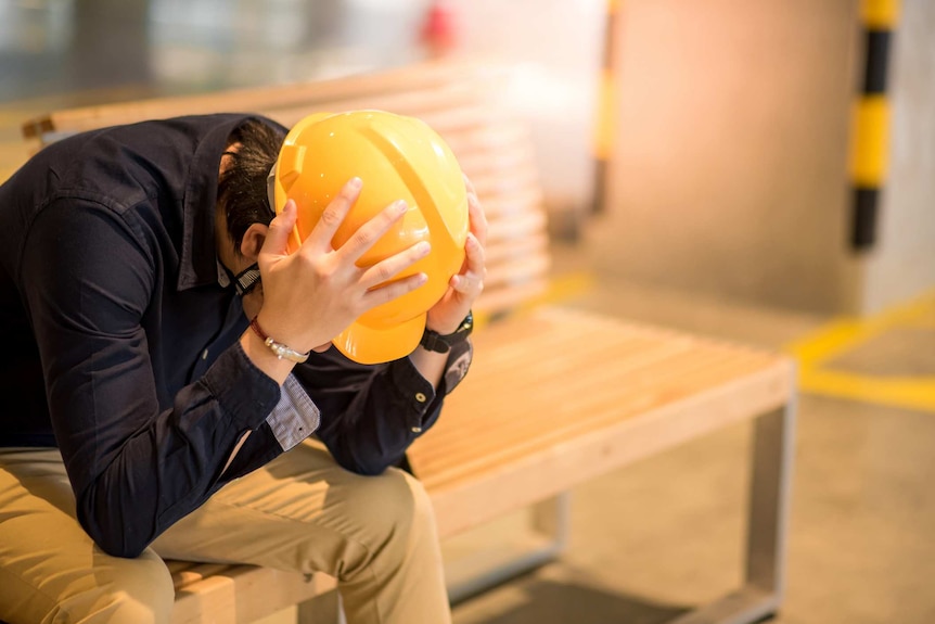Man wearing a hard helmet puts both hands on his head and leans over in a moment of stress and exhuastion.
