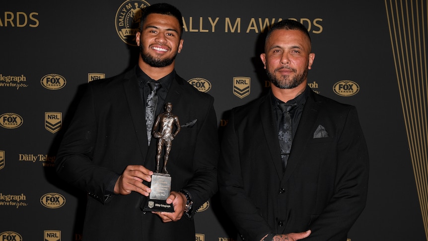 Two male adults, father and son, in black suits, posing for photographers at an award ceremony.
