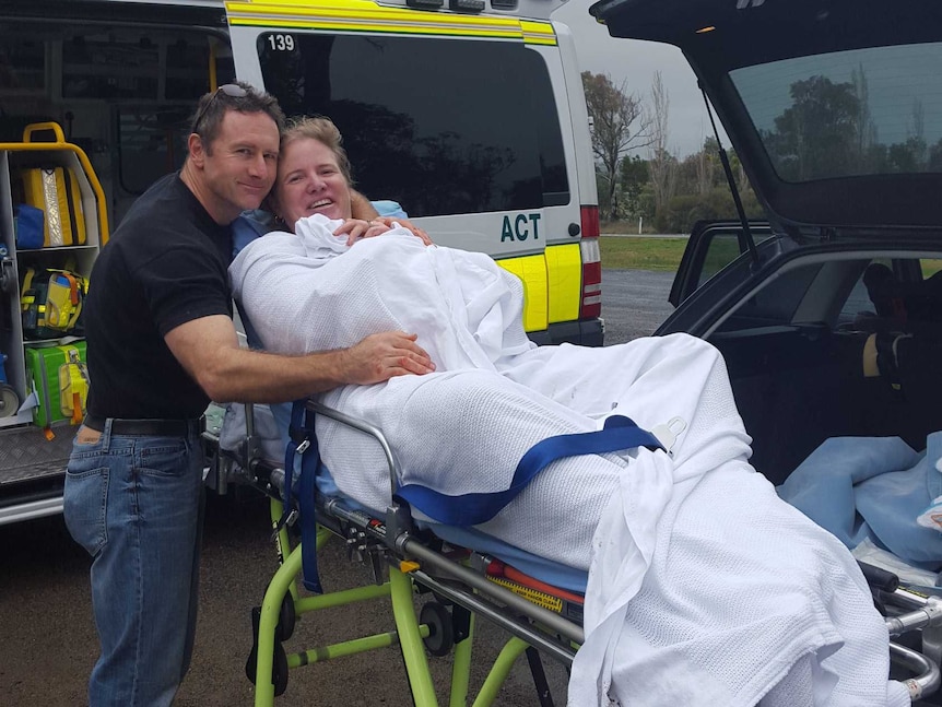 Woman on a stretcher bed in front of an ambulance at the side of the road holding a new born as her husband hugs her.