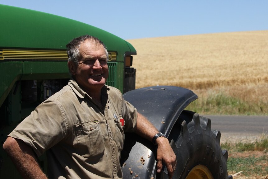 A man in a khaki shirt standing next to the tyre of a green tractor with a field of wheat in the background.