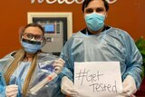 A woman and a man in full PPE hold a sign saying "swab for mob".