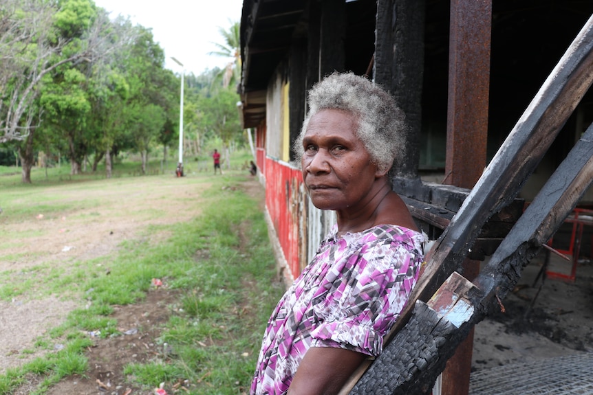 A woman leans against a burned building. She is looking into the camera with a stern expression.