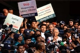 A crowd holding placards stand around a man who is holding his finger to his lips.