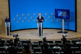 Jens Stoltenberg addresses a media conference from behind a grey lecturn on a timber stage, with a blue wall behind.