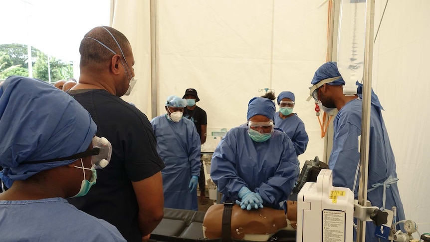 A person wearing scrubs demonstrates CPR on a dummy in front of other people dressed in blue scrubs and gloves.