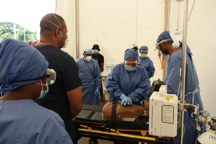 A person wearing scrubs demonstrates CPR on a dummy in front of other people dressed in blue scrubs and gloves.