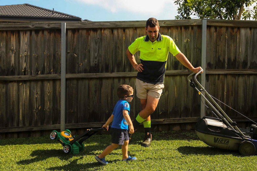 a young boy dragging a toy lawnmower next to his father who has a real lawnmower