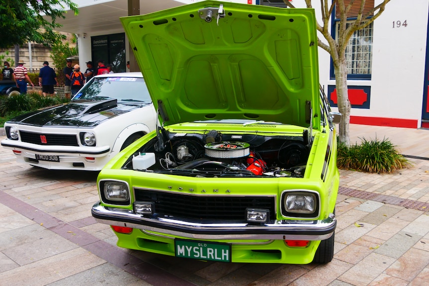 A green car sits on a pale street with its hood popped open.