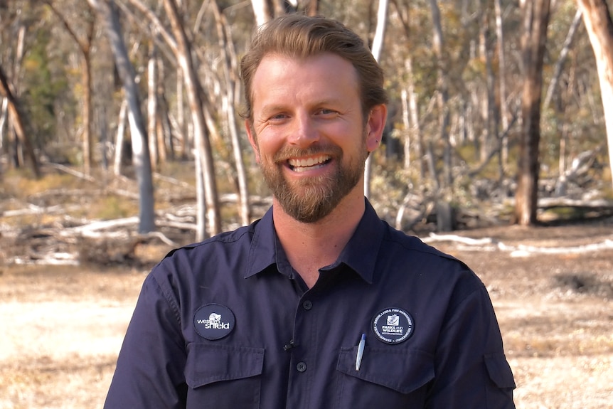 A man with dark blonde hair and a beard stands outside in bushland, with a big smile on his face.