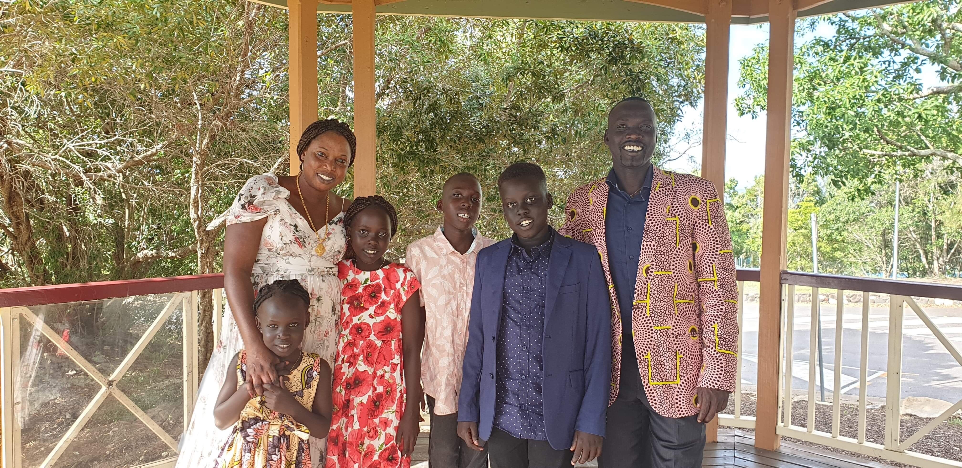 A woman, man and four children of different stand close together, smiling widely, wearing brightly coloured clothes.