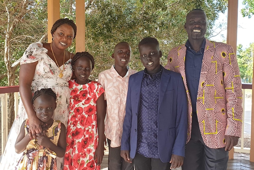 A woman, man and four children of different stand close together, smiling widely, wearing brightly coloured clothes.