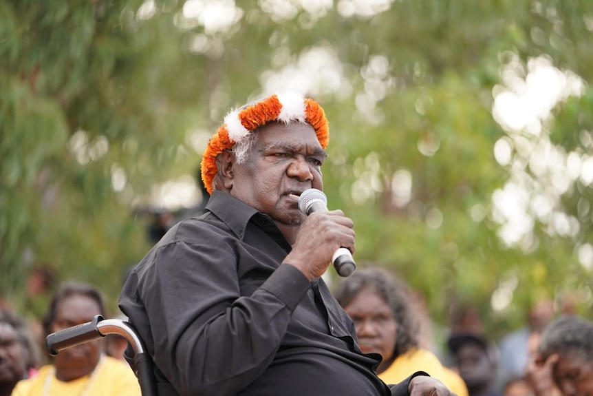 An older Aboriginal man wearing a black shirt and an orange and white headband speaks into a microphone.