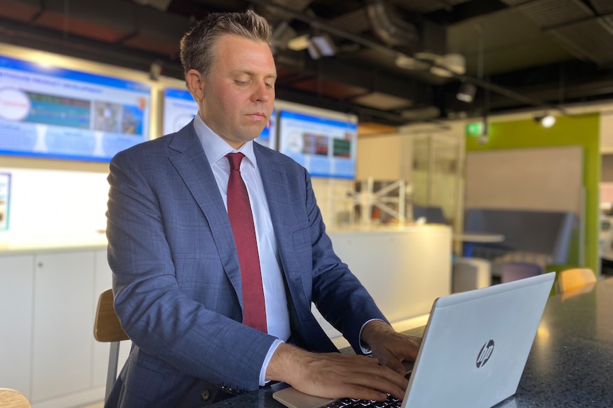 A middle aged Caucasian man wearing a blue suit, shirt and red tie sits at a bench working on a silver HP laptop.