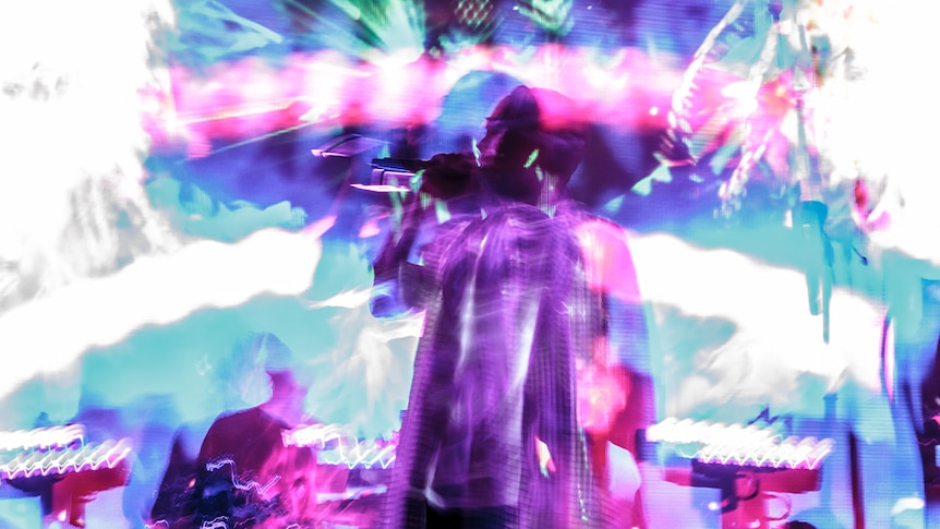The vocalist from Pnau stands on stage with kaleidoscopic, colourful lights surrounding them.