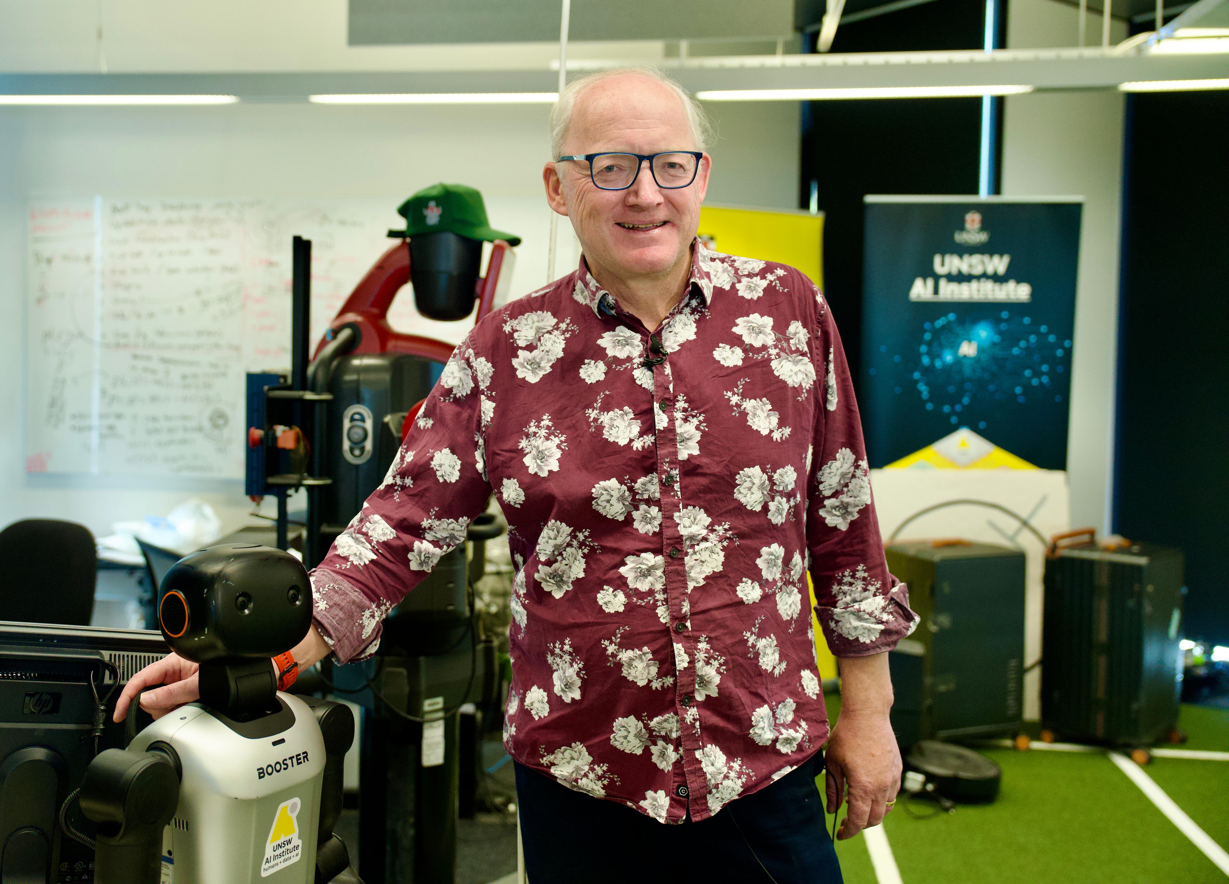 A grey haired man in black glasses smiles at the camera. He wears a patterned marron and white shirt.