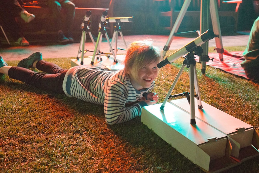 A young girl lays on the ground and looks through the telescope.