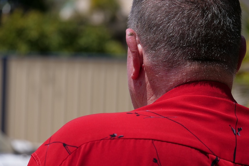 A photo of the back of an aged man's head. He has a sun spot on his ear and wears a red shirt.