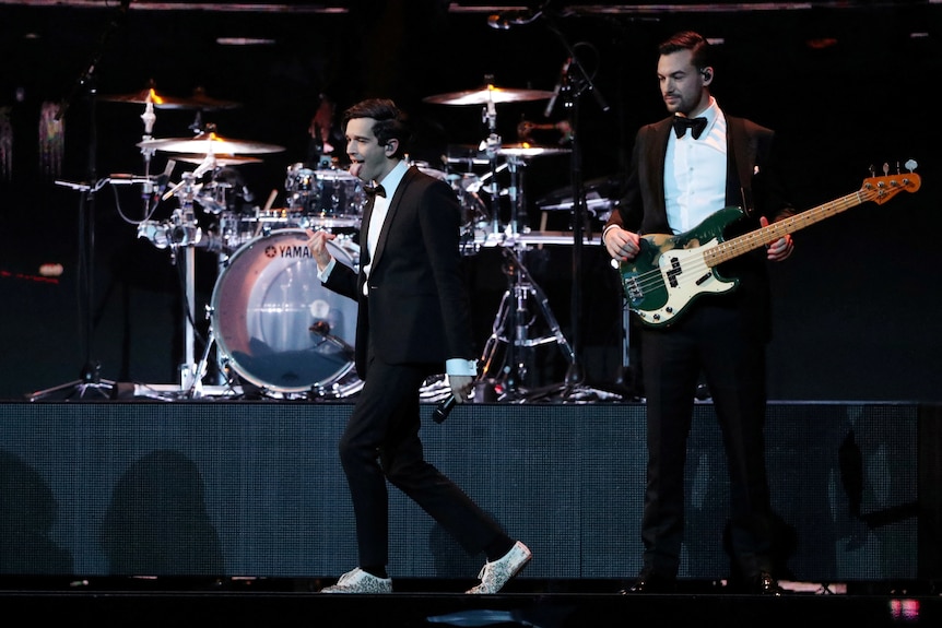 A lead singer and a guitarist, wearing tuxidos, perform on stage in front of a drum set.