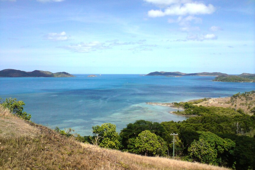 View over grassy hilltop to a wide, shallow bay with clear blue water and distant islands. 