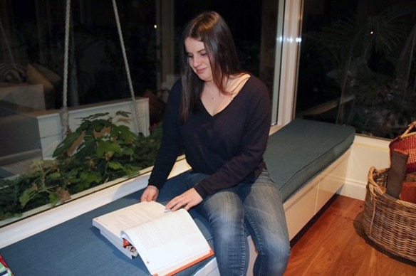 Ellie O'Grady sits on an indoor bench near a window, reading a book.