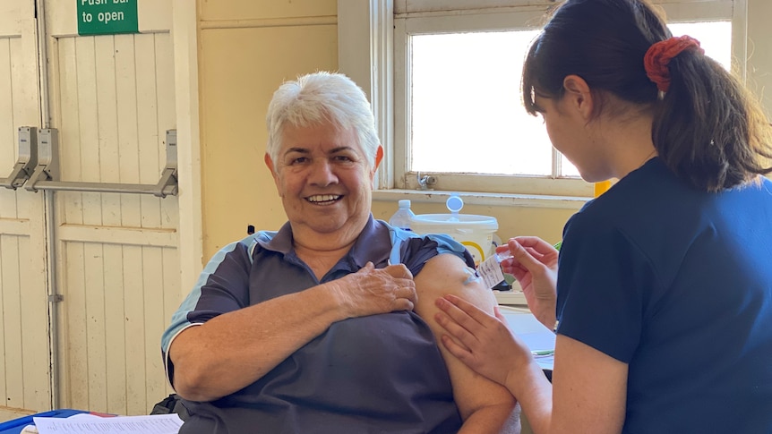 Silver-haired woman getting needle in her left arm from a dark-haired nurse, back to camera, in navy blue. They're in a hall.