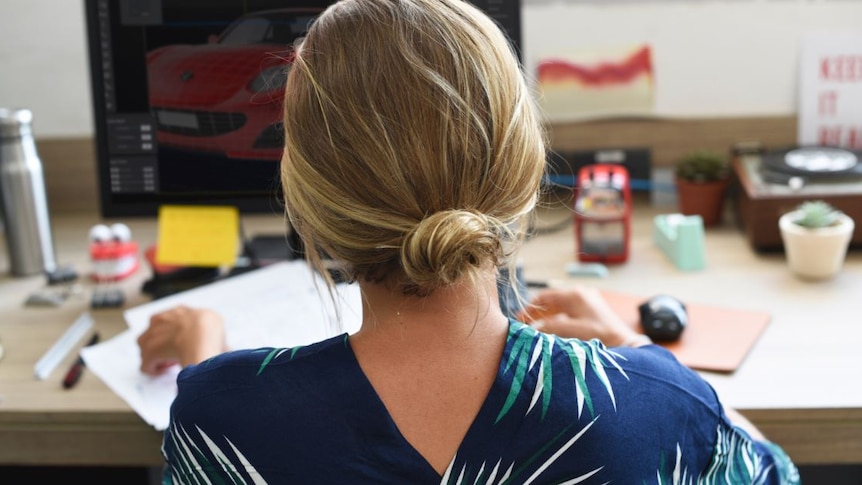 A woman with blonde hair sits at a computer desk inside an office.