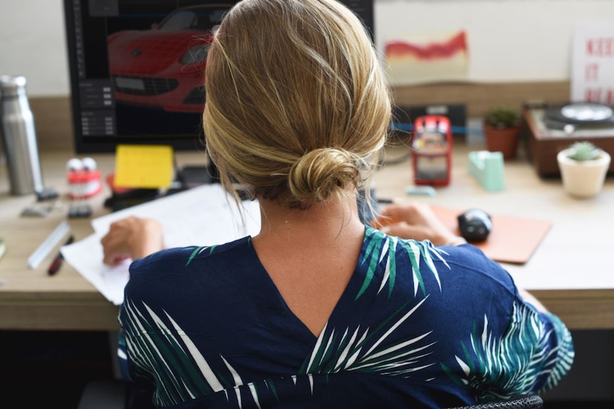 A woman with blonde hair sits at a computer desk inside an office.