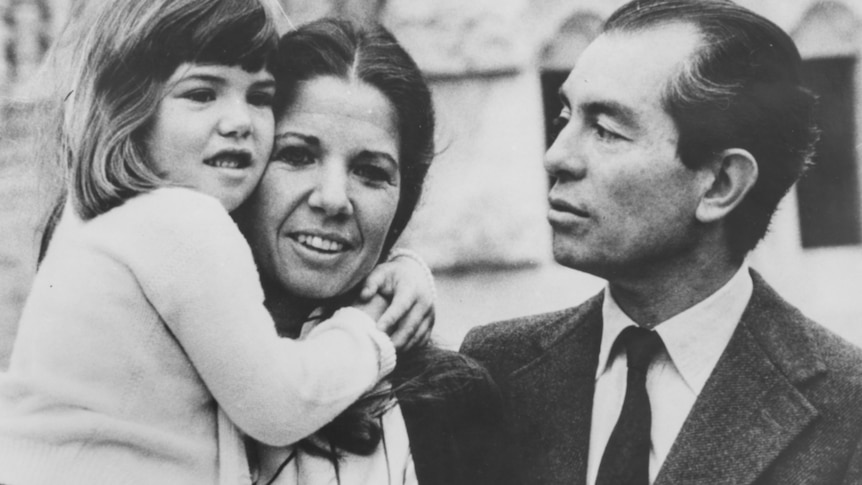 Black and white photo of a young girl in her mother’s arms, father looking on.