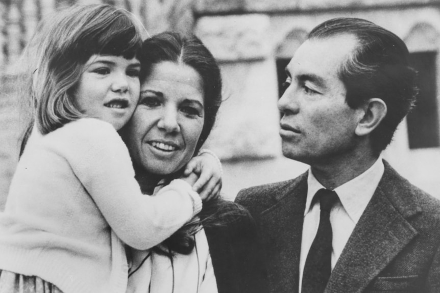 Black and white photo of a young girl in her mother’s arms, father looking on.