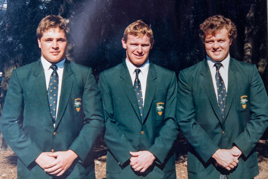 Three young men in green Australian rugby blazers stand with their hands in front of them looking at the camera.