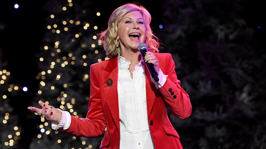 A woman in a red jacket holds her hand out as she sings.