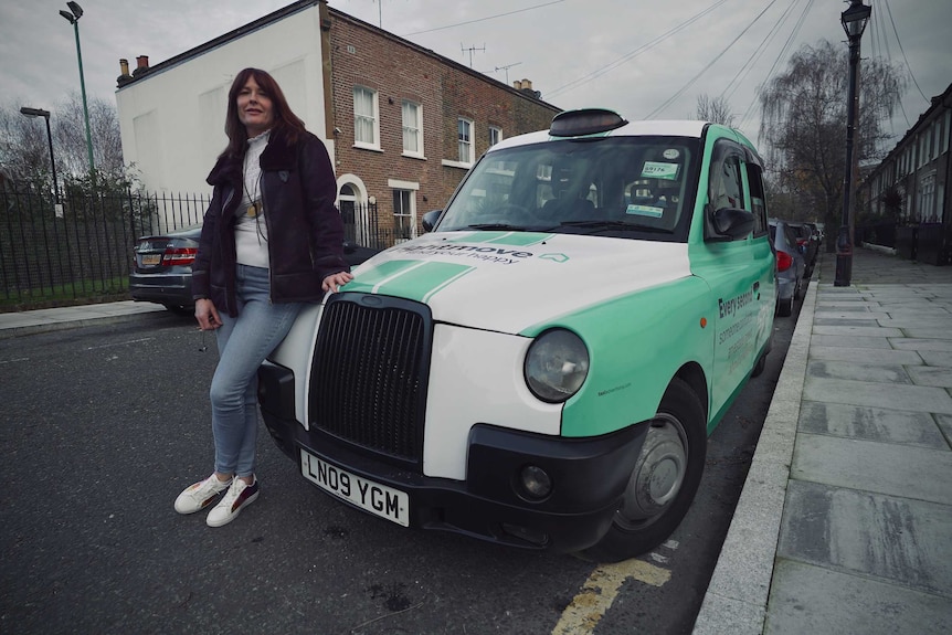 Sarah Tobias stands next to her black cab on a street in east London on an overcast day.