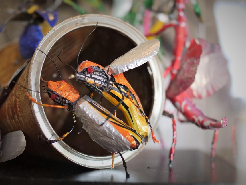 Two colourful insects made from aluminium cans, near the mouth of an overturned can.