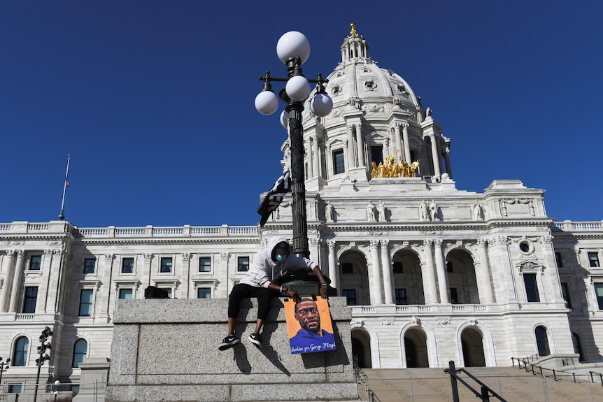 A black man in a hoodie sits on a platform with a poster in front of an ornate white building