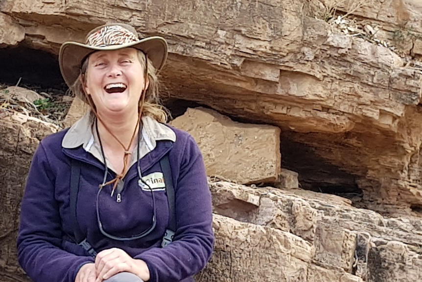 A laughing woman wearing a broad-brimmed hat surrounded by rocks with a cliff behind her