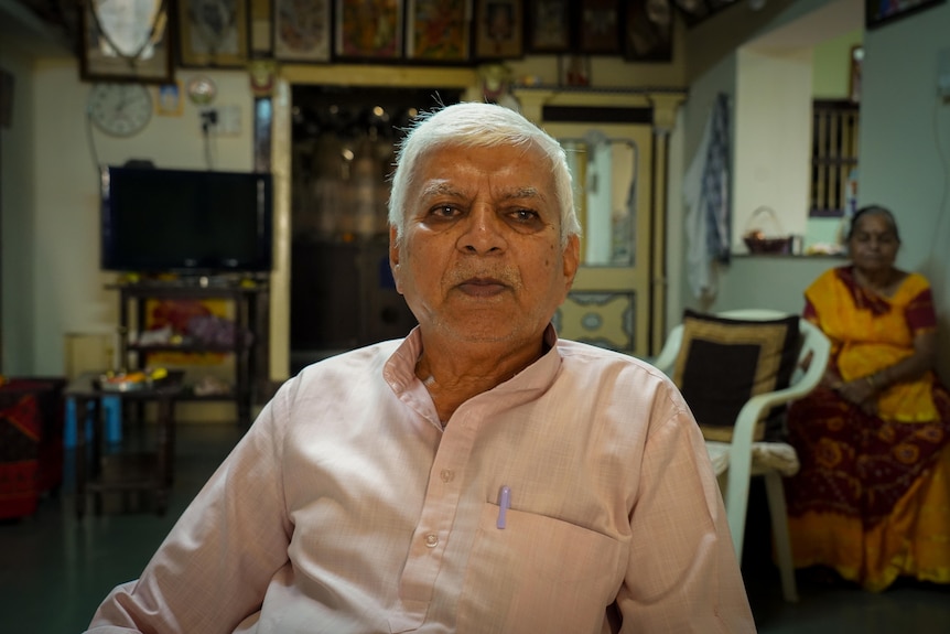 An older man in pale pink shirt with pen in pocket looks just beyond camera while a woman sits behinid