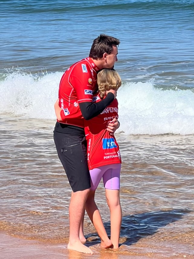 A man in a red rashie hugs a child on a beach with the swell behind them 