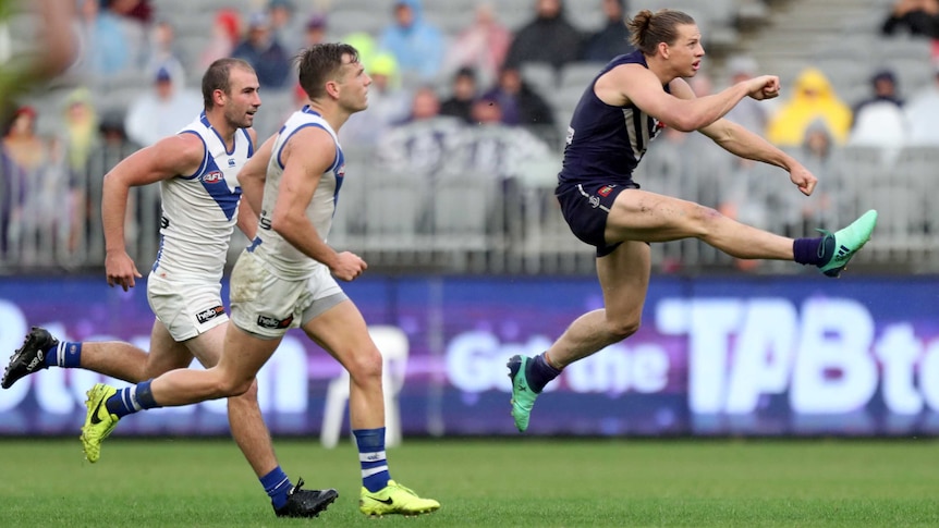 An AFL player is mid-air after kicking the ball clear, as defenders chase him.