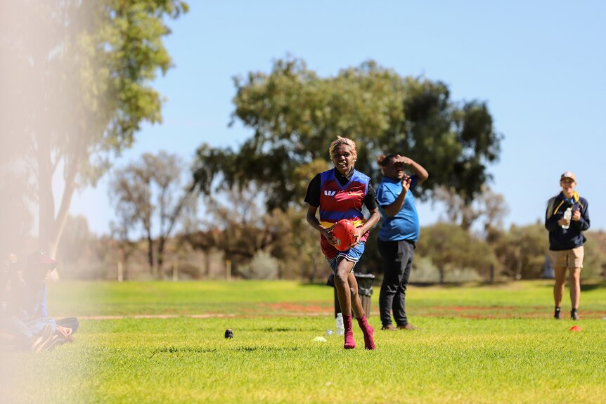 A young Aboriginal woman with a red football wearing a team uniform runs across a green oval
