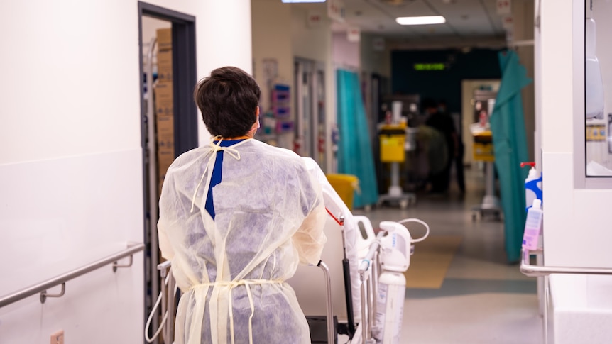 A nurse dressed in a pale yellow PPE gown is pictured from behind pushing a bed through the corridor of the Alfred Hospital.