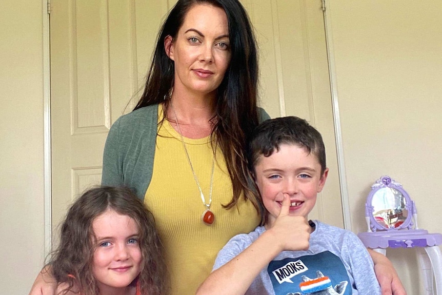 A woman facing the camera with her arms around her two young children.