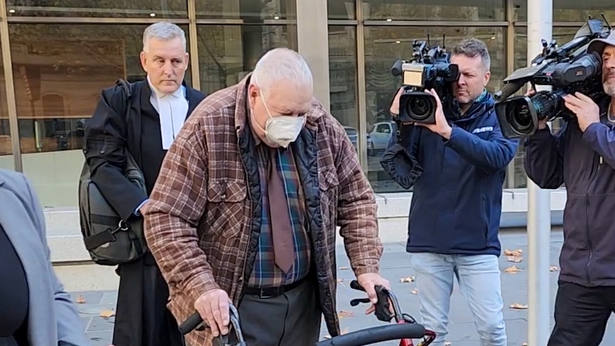 Hutchins wears a covid face mask and uses a walker as he walks past tv cameras outside court