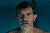 Antonio Banderas wears blue swimming trunks and stands with muted expression in pool, his head and shoulders above water.