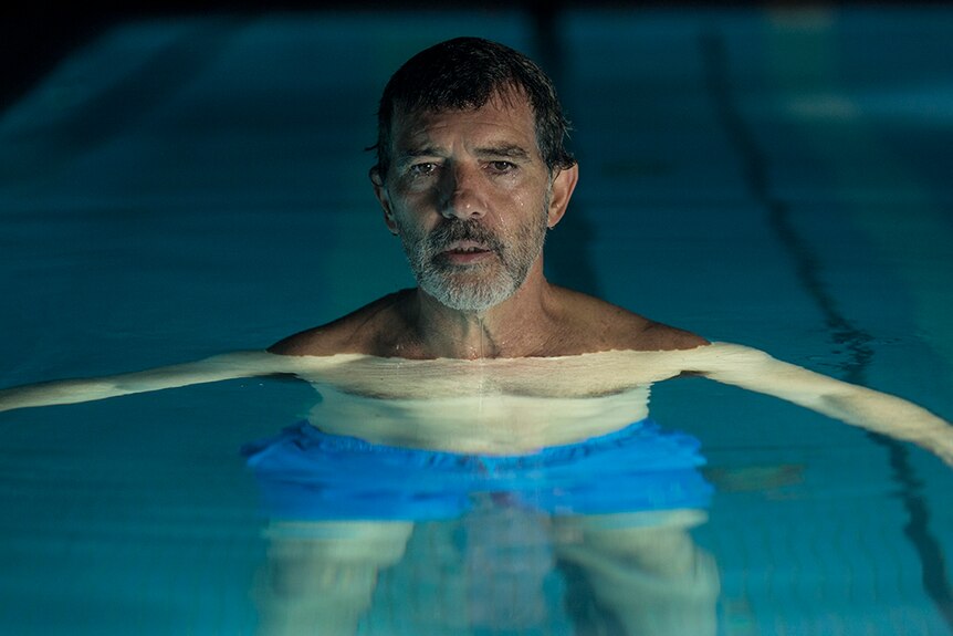 Antonio Banderas wears blue swimming trunks and stands with muted expression in pool, his head and shoulders above water.
