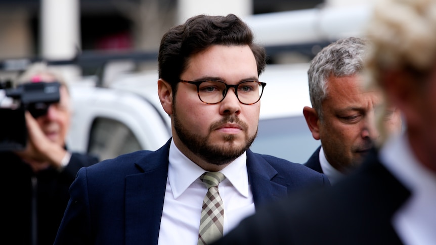 Bruce looks serious, wearing a suit and tie and glasses, as he walks surrounded by people, a video camera in the background.