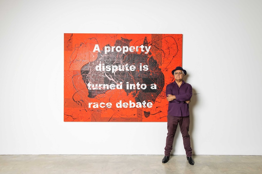 Richard Bell standing beside his artwork "A property dispute is turned into a race debate" (2019)